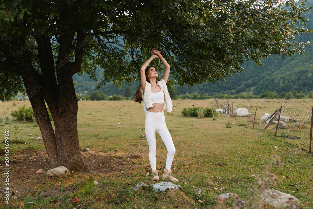 Naklejka premium Young woman in white croptop and pants dances under a large tree in a countryside field, radiating carefree joy and natural serenity.