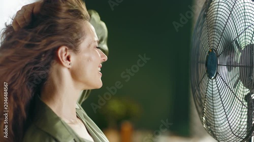 A happy woman with long brown hair, wearing a green shirt, experiences refreshing relief from summer heat as an electric fan blows her hair, conveying comfort and wellbeing.