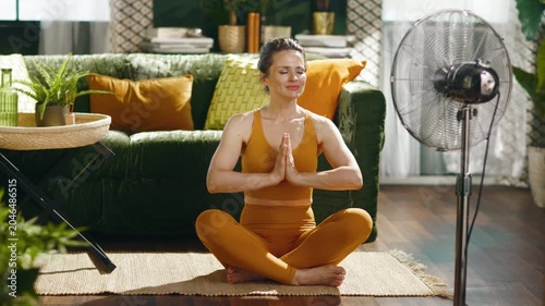 A serene woman in yellow sportswear practices yoga or meditation in a bright living room. A stand fan nearby provides comfort and heat relief during her mindful home wellness routine.