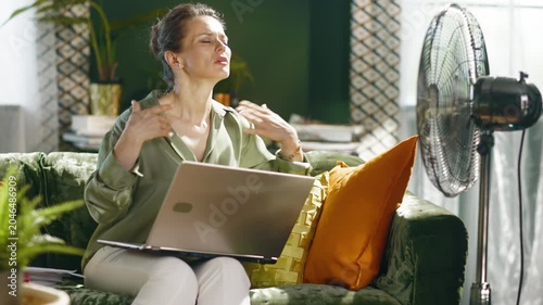 A middle-aged woman works on her laptop from a green velvet couch, fanning herself due to intense summer heat. A nearby fan tries to alleviate the challenging home office environment.