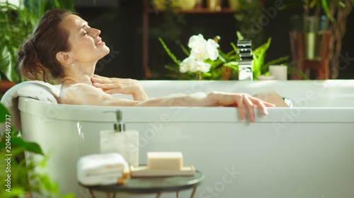 A serene woman with eyes closed gently touches her neck while relaxing in a luxurious bathtub. Surrounded by lush plants & soft natural lighting, she embodies tranquil self-care & indulgent pampering.