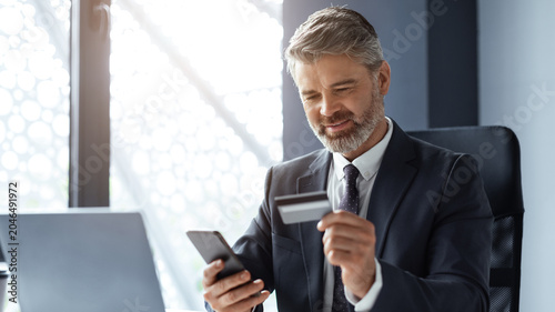 Quadro su tela Focused Middle Aged Caucasian Man Holding Credit Card While Using Smartphone At Desk In Modern Office