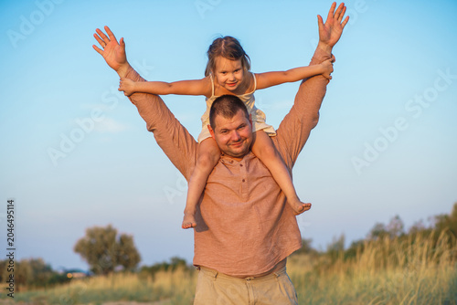 A father and his little daughter play and dance happily on a sandy lake beach at sunset. Happy childhood. The father put his daughter on his neck.