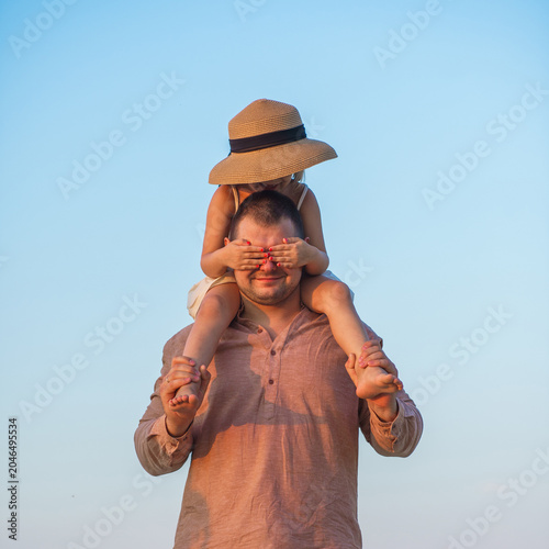 A father and his little daughter play and dance happily on a sandy lake beach at sunset. Happy childhood. The father put his daughter on his neck.