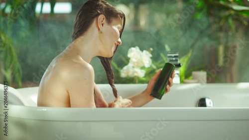 A radiant woman with wet brown hair smiles in a bubbly white bathtub, holding a green hair product & examining her hair. Lush plants & soft light create a serene, spa-like self-care experience.