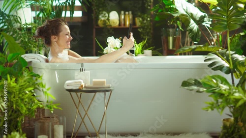 A smiling woman with a bun enjoys a luxurious bubble bath, using a smartphone. Surrounded by lush greenery & candles, capturing self-care, comfort & digital connection in a tranquil setting.
