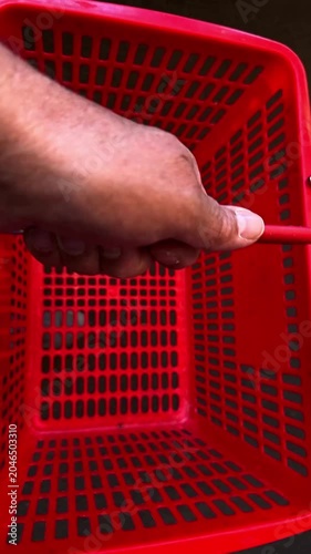Close-up view of a person's hand holding and carrying an empty red plastic shopping basket, getting ready to shop in a supermarket, slow motion