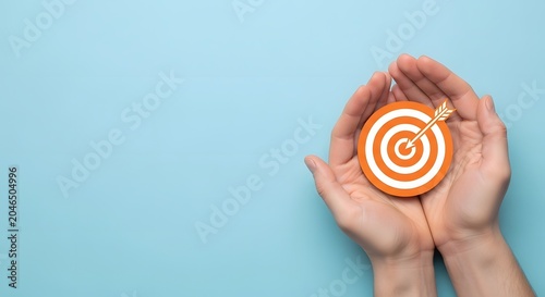 A pair of hands gently cupping and holding an orange and white target with an arrow hitting the bullseye, set against a light blue background, symbolizing goals and achievement.