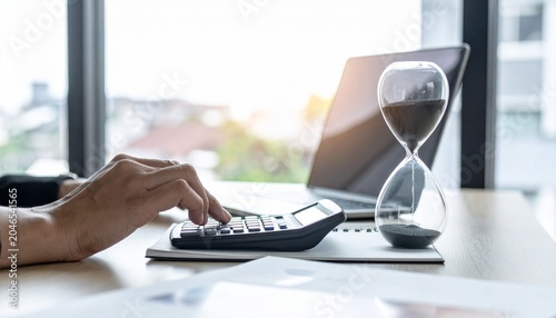 A close-up shot of a person's hands using a calculator next to a laptop and an hourglass, symbolizing the passage of time and financial management