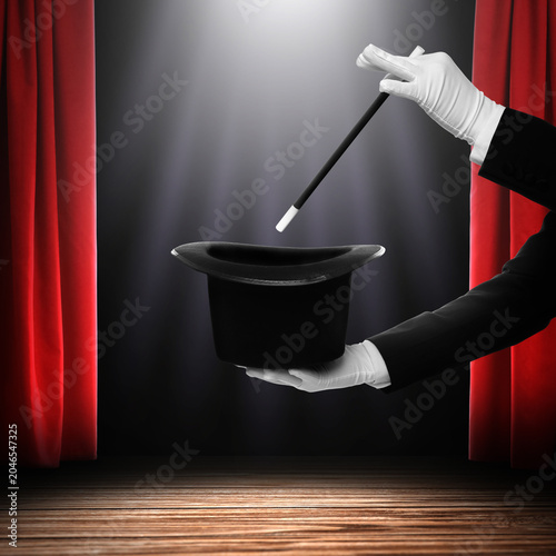 Photos Magician performing trick with wand and top hat on stage against red curtains, c
