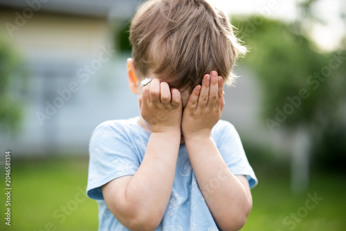 Tableau sur toile Shy little boy covering his face with hands while holding a daisy flower in a sunny garden
