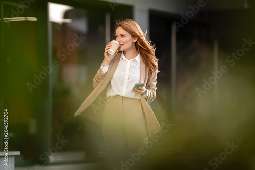 Obraz na plátně Businesswoman drinking coffee and using smartphone while walking near office bui