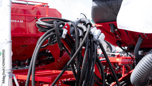 Detailed close-up of red agricultural machinery with bundled cables, hydraulic hoses, metal brackets, and mechanical components in a bright outdoor setting, showing industrial