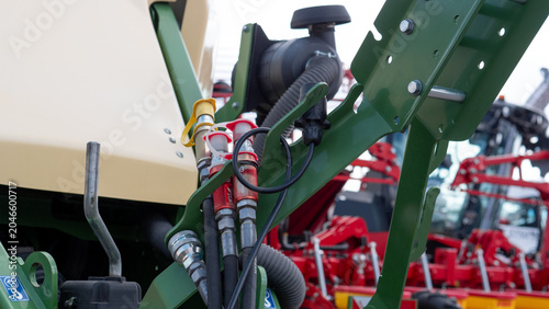 Detailed view of farm equipment showing green painted metal parts, hydraulic hoses, connectors, bolts, and mechanical linkages, with additional machinery blurred in the