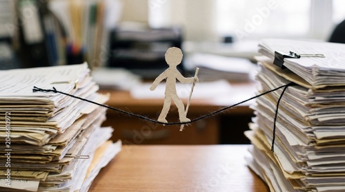 A paper figure uses a tiny pole to walk a thin black string stretched between towering stacks of files. It visualizes the precarious balancing act of a stressful workload.