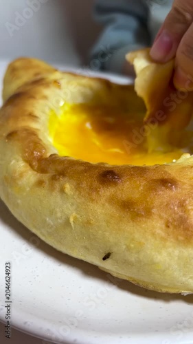 Vertical 4K video close up. Female hand of white woman breaks off piece of Adjarian style khachapuri and dips it into yellow yolk of an egg. Concept of food, national Georgian cuisine, cafe, eat, diet