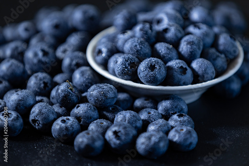 Fresh raw blueberries with water drops scattered around a white bowl on a dark background.
