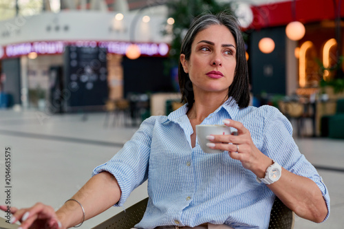 Mature woman with graying hair sitting in a casual striped shirt, holding a coffee cup and looking away, contemplating thoughts in a bright indoor cafe area