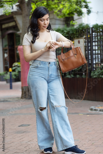 Woman walking on brick sidewalk near shops, adjusting wristwatch and holding brown leather handbag