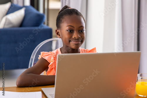 Youth African American female sitting at wooden table using silver laptop, coral top, bun, juice