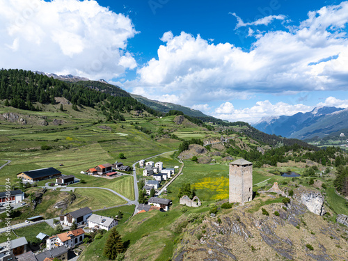 Aerial view of the historic Chaste Steinsberg tower ruins overlooking the village of Scuol and lush green alpine meadows under a cloudy blue sky Scuol, Graubunden, Switzerland.