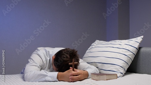 Tablou pe pânză Man kneeling in bed with head bowed, holding a wooden cross and spiritual holy b