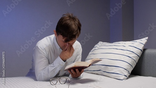 Vászonkép Young man reading in bed late at night, rubbing sore eyes from fatigue and blue