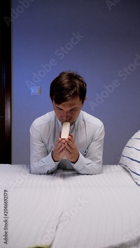 Fotografie Man kneeling on a bed in a quiet bedroom, holding a bible to his lips in prayerf
