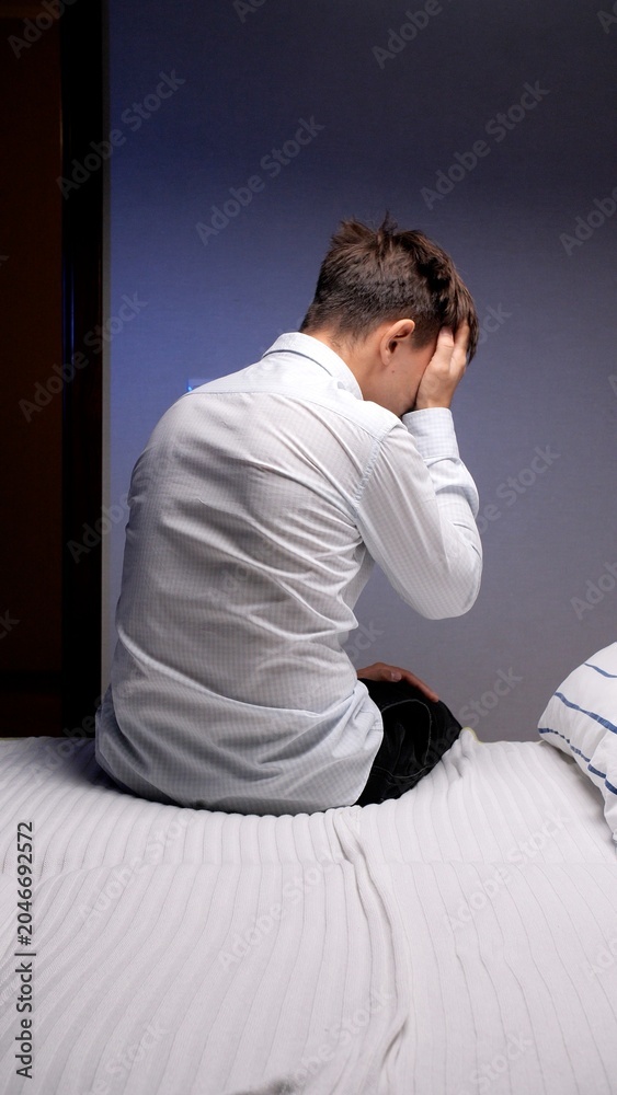 Fototapeta premium Young adult man sitting on the edge of a bed, covering his face with hands, feeling distressed and overwhelmed by sadness, depression, or a headache late at night