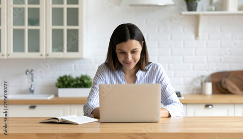 Focused woman working on a laptop at a wooden counter in a modern kitchen, natural light, casual work from home, domestic scene