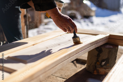 Close up of man hand applying clear varnish with brush on spruce wood door outdoors in winter