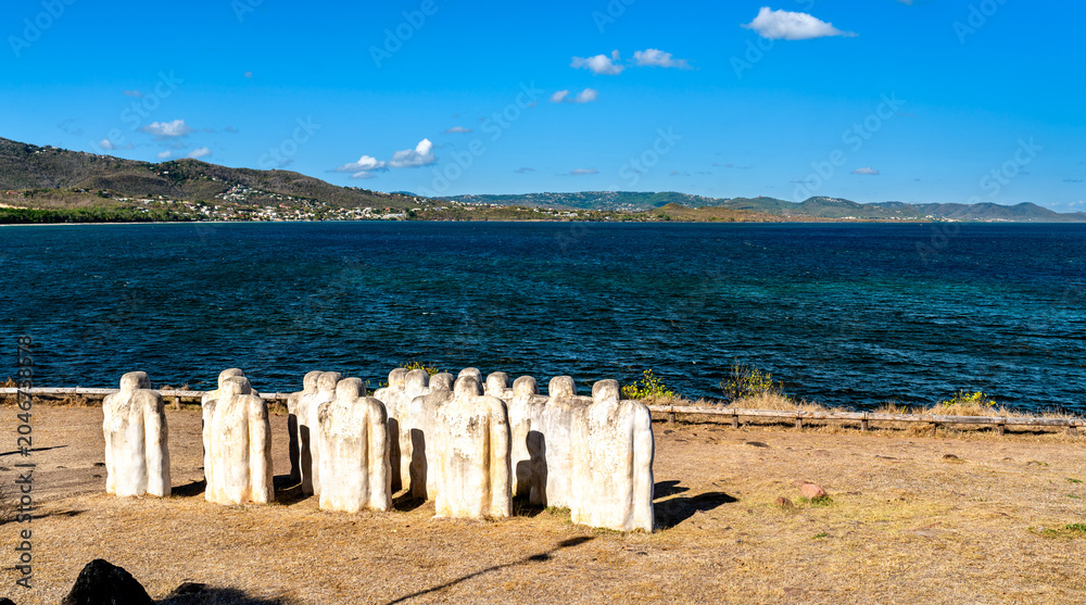 Obraz premium The Cap 110 Memorial at Anse Caffard in Martinique features white stone statues overlooking the blue Caribbean Sea and coastal hills.