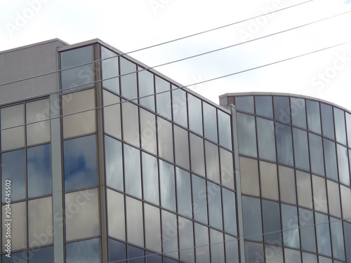 The glass window facade of a modern office building reflects a cloudy sky behind overhead power lines.