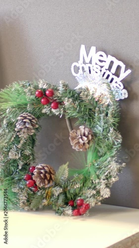 Vertical video: Panning camera revealing square wreath on tabletop and monitor, Merry sign blurring