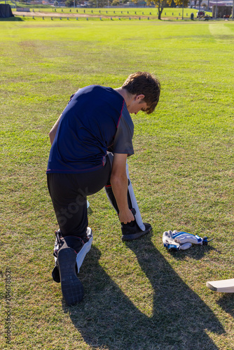 Male athlete is kneeling on grassy sports field fastening cricket leg pad with gloves nearby