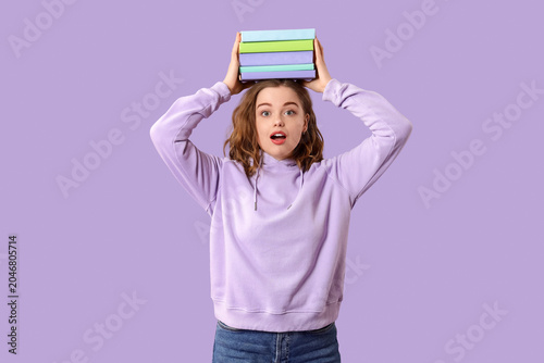 Surprised young woman with stack of books on her head against lilac background