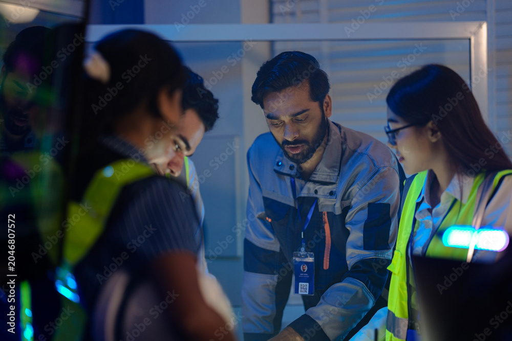 Obraz premium An Indian supervisor leads a night shift briefing in a smart factory control room. He discusses an AI automation system or incident response with his female engineers.