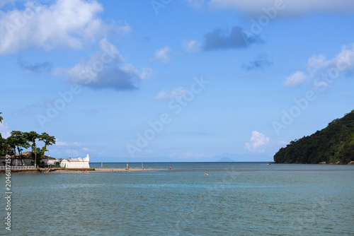 View of Bertioga channel entrance featuring historic fort, calm sea, palm trees, and coastal landscape under blue sky in Brazil.