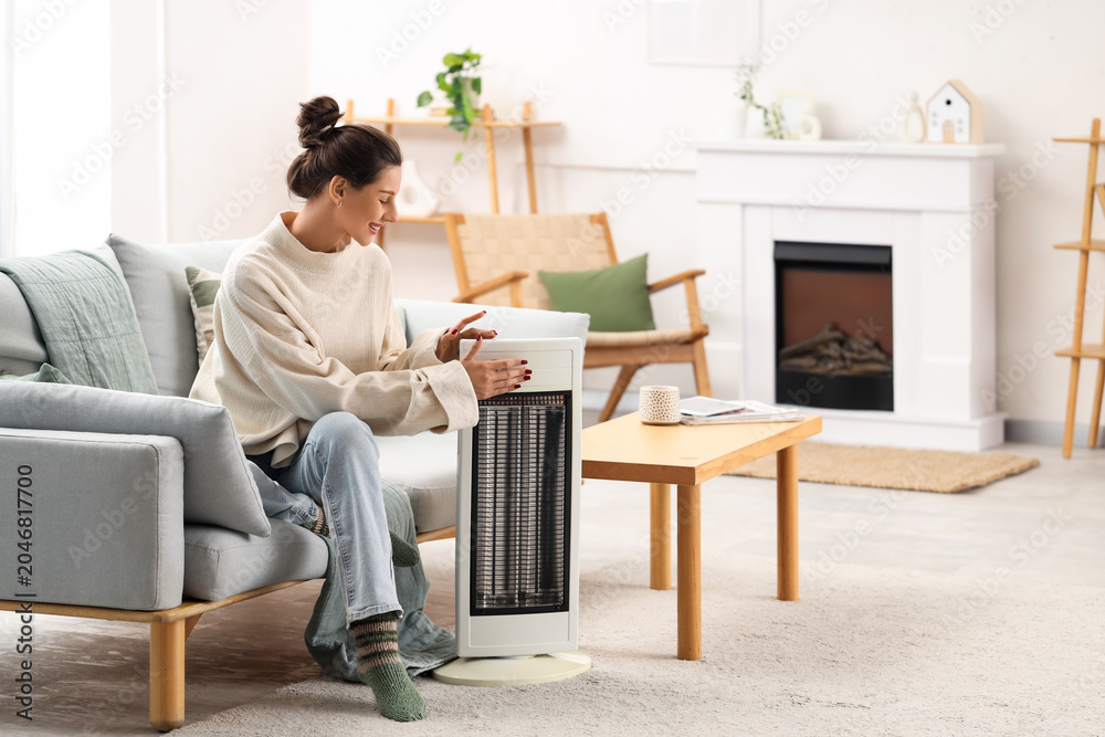 Fototapeta premium Young woman with electric heater sitting on sofa at home