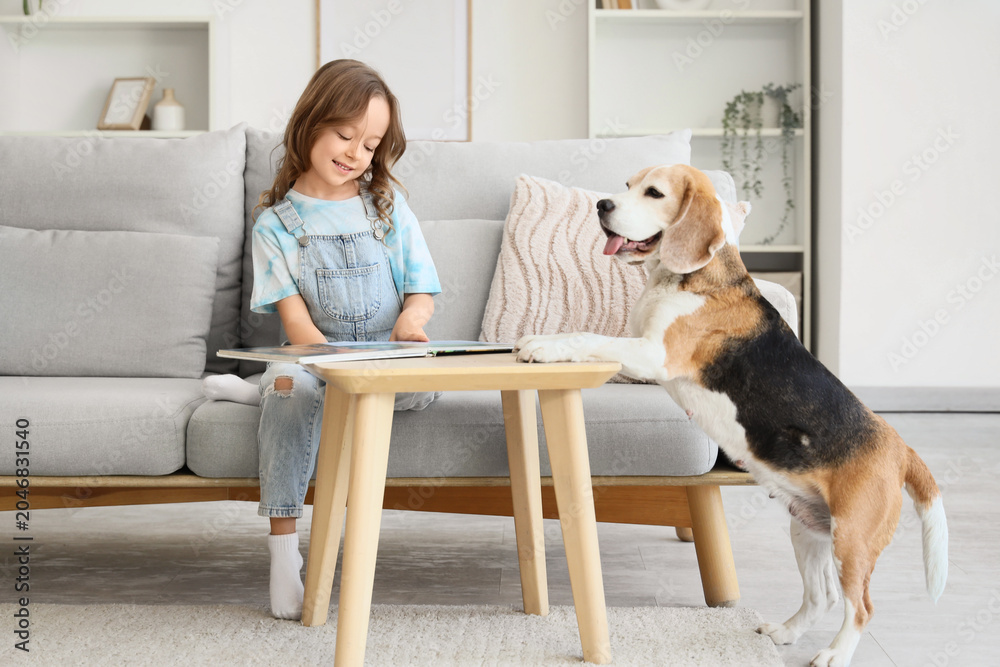 Naklejka premium Cute little girl with Beagle dog reading book at table in living room