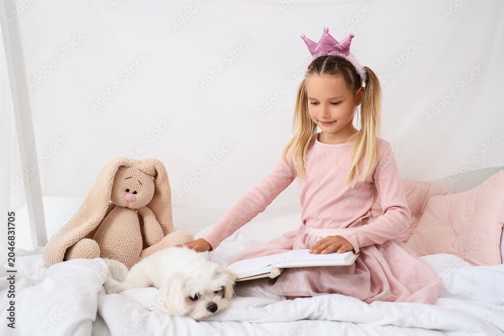 Naklejka premium Cute little happy girl with Maltese dog and toy reading book on bed at home