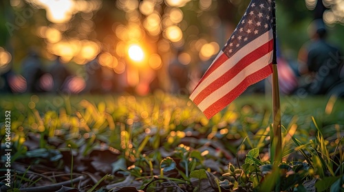 Against the soft golden glow of sunset in an outdoor autumn setting, a small American flag stands planted in grass-dusted ground as the sole sharp focal point, with warm circular bokeh from sunlight