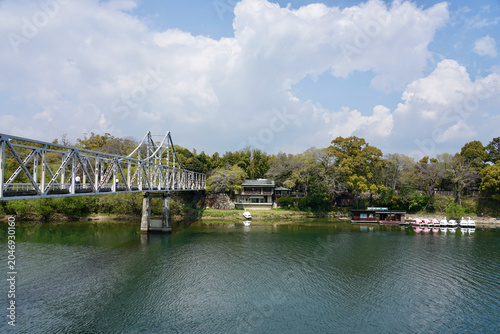 Steel Truss Pedestrian Bridge Over Calm River With Trees, Boat and Cloudy Sky