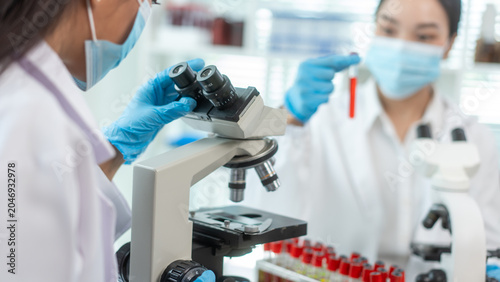 Scientists in medical masks and gloves work with microscopes and blood test tubes in a bright laboratory, showing teamwork, research, diagnosis, and healthcare science.