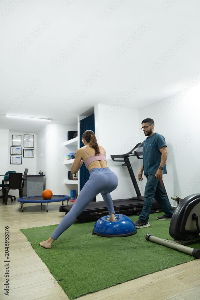 Fototapeta premium Female patient practicing balance and lower body stability on a BOSU trainer while being monitored by a physiotherapist during rehabilitation therapy.