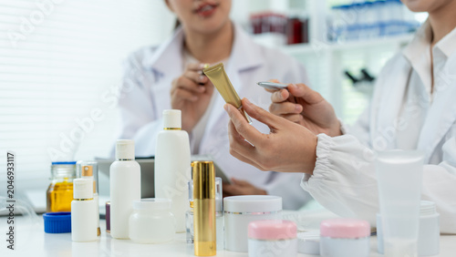 Two female scientists in white lab coats test skincare products at a laboratory table with bottles, jars and a laptop, showing research, teamwork and cosmetic development.