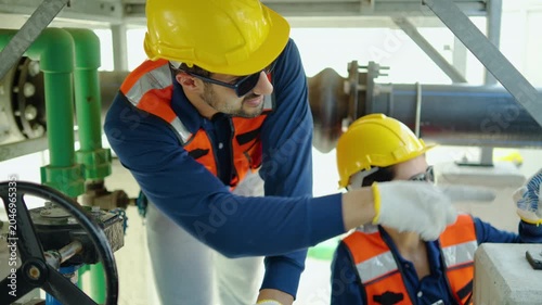 Two professional industrial engineers wearing protective safety gear carefully inspecting pipeline valves and discussing maintenance data on a digital tablet inside a large manufacturing plant.