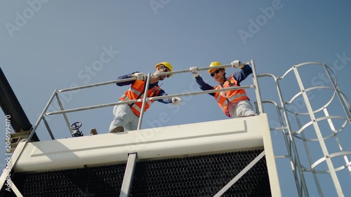 Two professional male industrial engineers wearing safety vests and hard hats stand on a commercial cooling tower platform against a clear blue sky, inspecting the HVAC systems and discussing work.