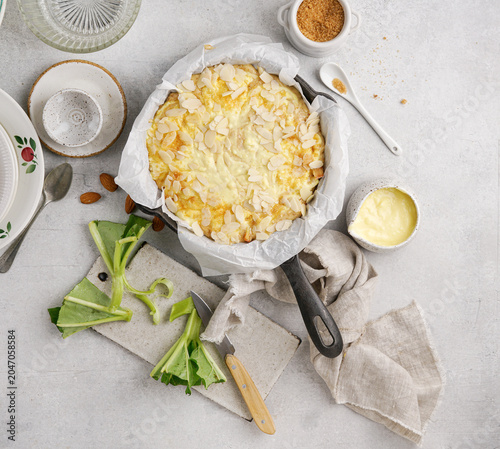 Top view of unbaked English rhubarb pudding with custard and almond flakes in cast iron skillet ready to bake on white concrete background flat lay