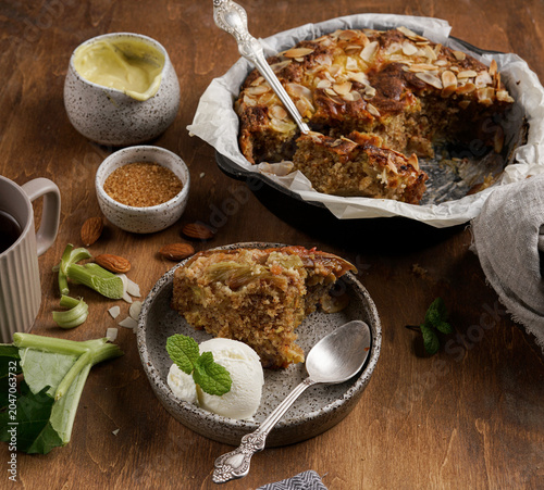 Side view of freshly baked English rhubarb pudding slice served with vanilla ice cream and mint on plate, cake in cast iron skillet on rustic wooden table background